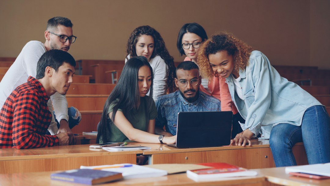 University students collaborating around a laptop in a lecture hall; photograph by Vitaly Gariev on Unsplash.